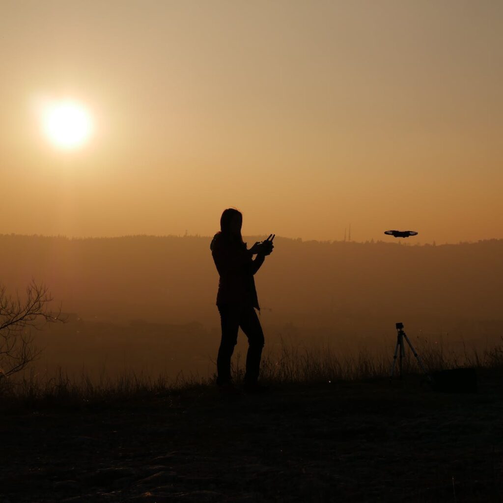 Full body silhouette of anonymous person standing on slope at sunset and using remote control for operating drone